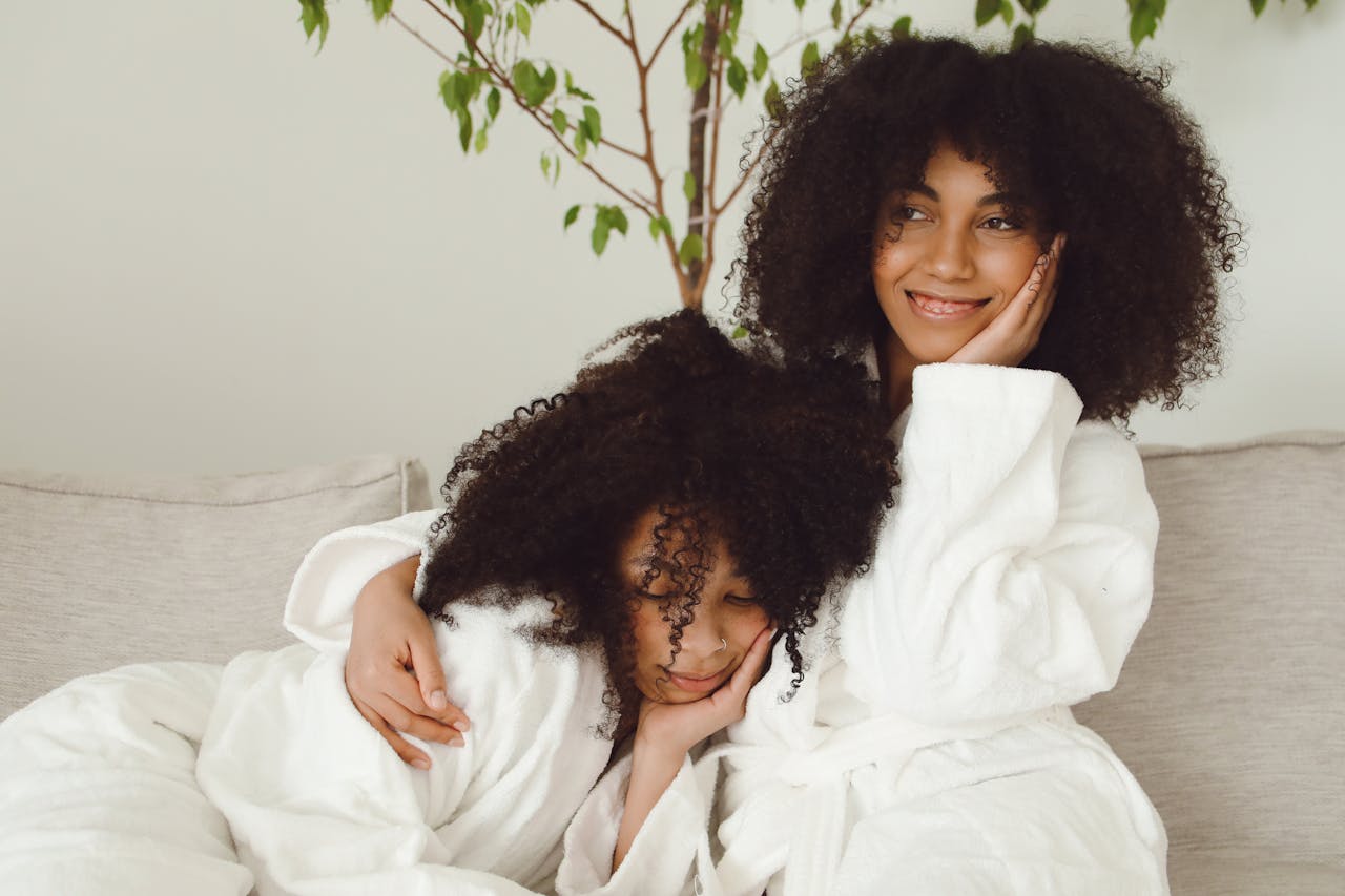 Two smiling women in bathrobes enjoying a relaxed moment on a sofa with lush green plant nearby.