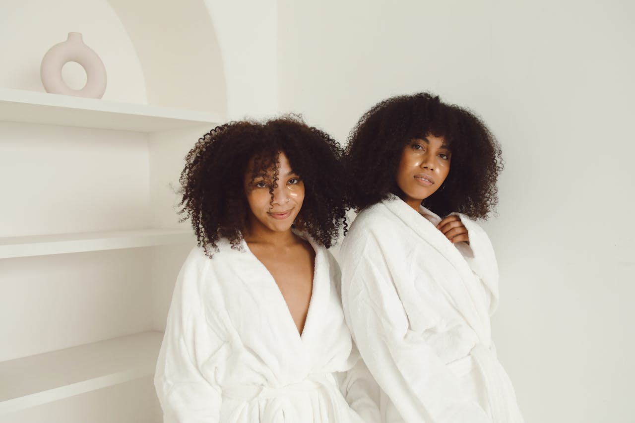 Two women with afro hair wearing white bathrobes posing indoors.