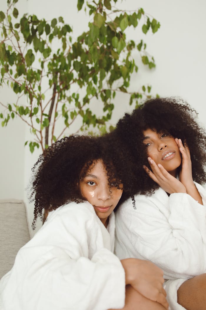 Two sisters with afro-textured hair in bathrobes share a tender moment indoors.