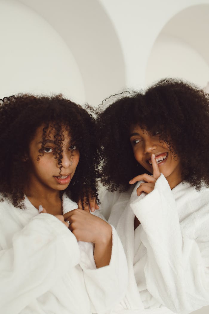 Two sisters with curly hair smiling warmly in white bathrobes indoors.