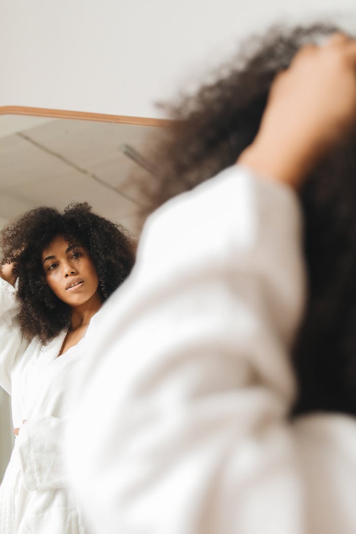 Woman with curly hair in a bathrobe looking at her reflection in a mirror indoors.
