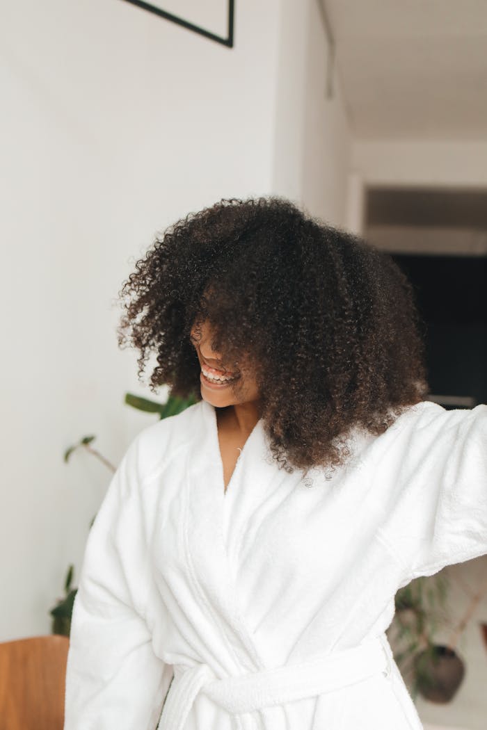 Smiling woman with curly hair wearing a bathrobe, enjoying a relaxed moment at home.